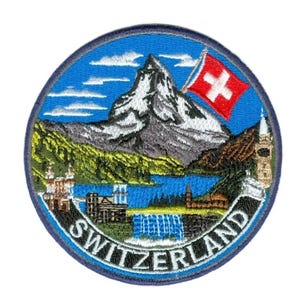 Op de afbeelding: Geborduurde patch met een Zwitsers landschap met een besneeuwde berg, een meer en een waterval. De Zwitserse vlag is zichtbaar. Het woord "SWITZERLAND" is wit geborduurd op een blauwe rand.
