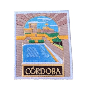 Könnte beinhalten: Gestickter Aufnäher mit beigefarbenem Hintergrund, der ein stilisiertes Bild eines Gebäudes mit einem Pool davor zeigt. Der Text "CÓRDOBA" ist in goldenen Buchstaben am unteren Rand des Aufnähers geschrieben.