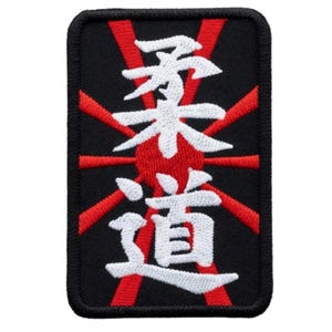 Puede incluir: Parche bordado con caracteres japoneses de Judo en blanco sobre fondo negro. Rayos rojos emanan de un círculo rojo central. El parche tiene un borde negro, creando un diseño llamativo.