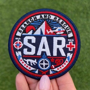 Könnte beinhalten: Ein kreisförmiger Aufnäher mit marineblauem Hintergrund und rotem und weißem Rand. Der Aufnäher zeigt den Text "SEARCH AND RESCUE" in Weiß, eine Berg-Grafik, einen Hubschrauber und die Buchstaben "SAR" in Weiß. Zusätzliche Grafiken sind ein Kompass und ein rotes Kreuz.