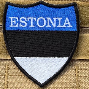 May include: A blue, black, and white embroidered patch in the shape of a shield with the word "ESTONIA" in white lettering. The patch has a hook and loop backing.