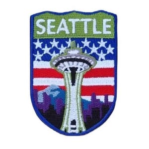 May include: Embroidered patch with the word "SEATTLE" above a depiction of the Space Needle. The design incorporates the American flag with red and white stripes and a blue field of stars. A purple cityscape is at the bottom.