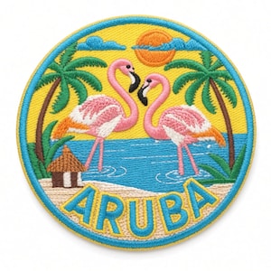 Op de afbeelding: Een ronde geborduurde patch met twee roze flamingo's die in blauw water op een zandstrand staan, met groene palmbomen, een kleine bruine hut en een oranje zon tegen een gele lucht. Het woord ARUBA is onderaan in het blauw geborduurd.