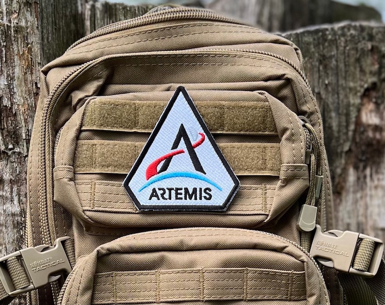 May include: A tan tactical backpack with a white triangular patch that says "ARTEMIS" in black letters. The patch has a red and blue graphic of a stylized letter "A" with a curved line.