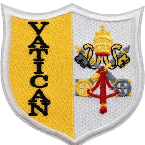 Può includere: Toppa a forma di scudo con la scritta "VATICAN" in lettere maiuscole nere su sfondo giallo. Il lato destro presenta uno sfondo bianco con un emblema dettagliato in giallo, rosso e grigio.