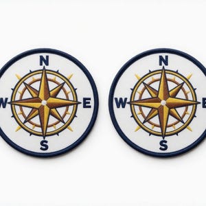 Op de afbeelding: Twee ronde patches met een kompasroosontwerp. De kompasroos is in goud- en bruintinten, tegen een witte achtergrond. De patches hebben een marineblauwe rand en de windrichtingen zijn aangegeven.