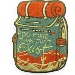 Può includere: Una toppa ricamata con un disegno di uno zaino da cartone animato con il testo "Do More Than Just Exist". Lo zaino è verde e arancione con un paesaggio desertico sullo sfondo.