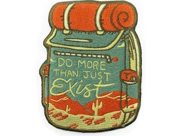 Pode incluir: Um remendo bordado com um desenho de mochila de desenho animado com o texto "Do More Than Just Exist". A mochila é verde e laranja com uma paisagem desértica no fundo.