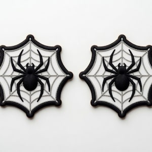 Peut inclure: Deux écussons en forme de toile d'araignée noire avec une araignée noire au centre. Les toiles sont blanches avec des lignes grises et les bords sont noirs. Parfait pour Halloween.