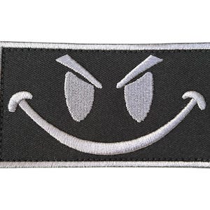 Könnte beinhalten: Ein schwarzer gestickter Aufnäher mit einem weißen Smiley-Gesicht mit wütenden Augen.