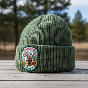 May include: An olive green ribbed knit beanie featuring a circular embroidered patch on the cuff. The patch displays a brown moose, blue mountains, and green pine trees with the text CANADA YUKON NATIONAL PARKS in red and white.