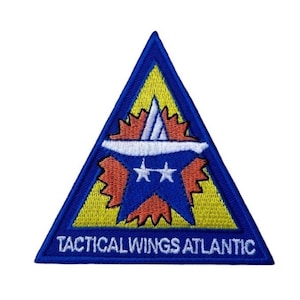Puede incluir: Un parche triangular azul con fondo amarillo. El parche presenta una estrella blanca con dos estrellas blancas más pequeñas en su interior. La estrella está rodeada por un diseño de rayos de sol rojo y naranja. El texto "Tactical Wings Atlantic" está impreso en blanco en la parte inferior del parche.