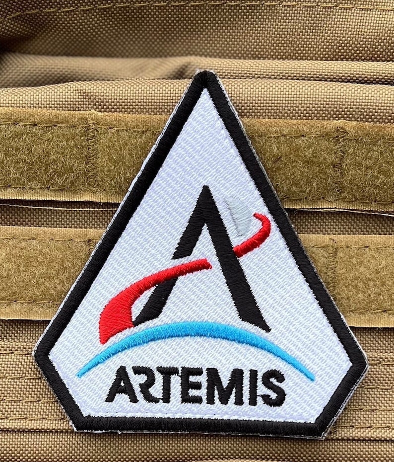 May include: White triangular patch with a black letter A and a red curved line. The patch has a blue curved line below the letter A and the word "ARTEMIS" in black letters below the blue line.