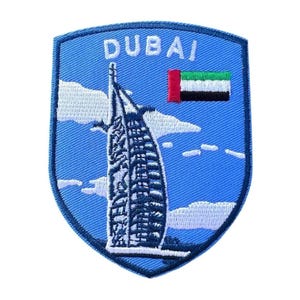 Op de afbeelding: Een blauwe geborduurde patch met het woord "DUBAI" en een witte omtrek van het Burj Al Arab hotel. De patch bevat ook een witte wolk en de vlag van de Verenigde Arabische Emiraten.