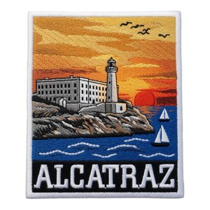 May include: A rectangular embroidered patch featuring a scenic view of Alcatraz Island with a lighthouse and building on a rocky cliff. The scene includes a sunset with orange and yellow hues, blue water with two sailboats, and the text ALCATRAZ.