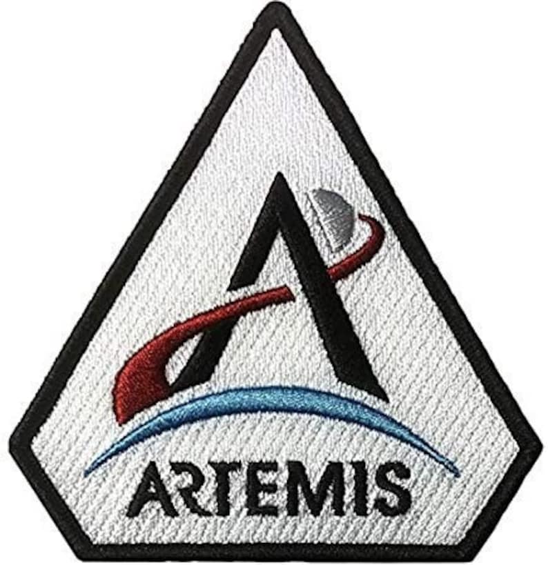 May include: A white triangular patch with a black outline and a stylized letter A with a red and blue orbit around it. The text "ARTEMIS" is printed in black at the bottom of the patch.