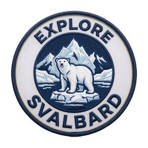 Op de afbeelding: Een ronde geborduurde patch met een witte ijsbeer die op een ijsschots staat. De achtergrond toont blauwe en witte bergen. De tekst EXPLORE staat in een boog bovenaan en SVALBARD onderaan in donkerblauwe letters.