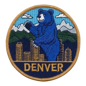 Puede incluir: Parche bordado circular con un oso azul sobre un fondo de montañas y ciudad. La palabra "DENVER" está escrita en letras doradas. El parche tiene un borde dorado. El oso es el foco central del diseño.