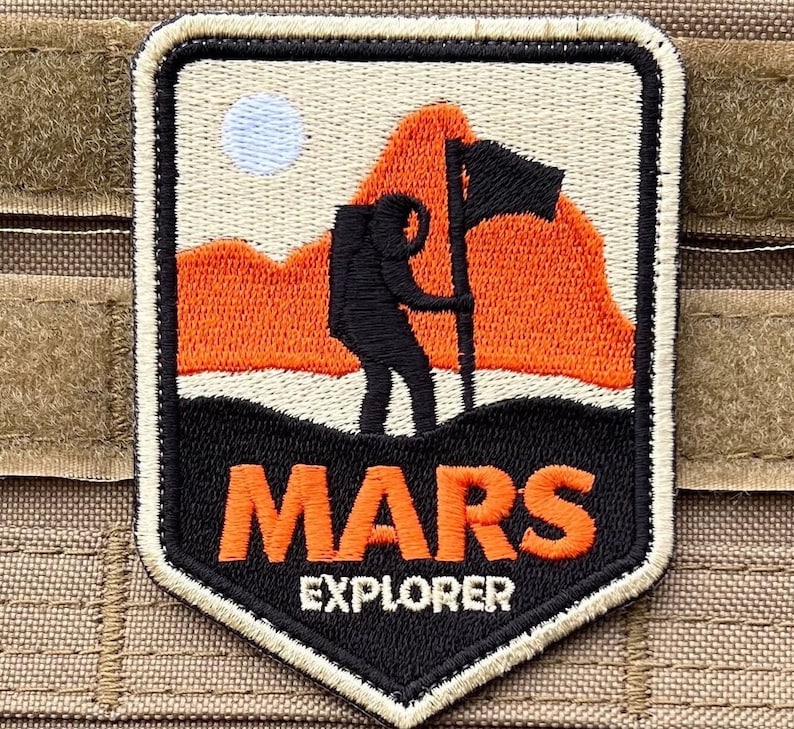 May include: A black and orange embroidered patch featuring a silhouette of a person holding a flag on a mountain. The text "MARS EXPLORER" is below the image.