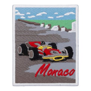 Op de afbeelding: Geborduurde patch met een rode, gele en zwarte raceauto op een grijze baan, met een blauwe lucht en groene heuvels op de achtergrond. Het woord "Monaco" is in rode letters geschreven. De patch heeft een witte rand.