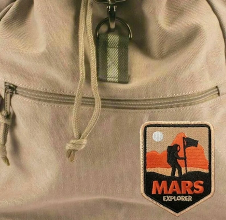 May include: A tan canvas backpack with a zipped pocket and a brown patch featuring an orange and black illustration of a person standing on a red planet with a flag. The patch reads "MARS EXPLORER".