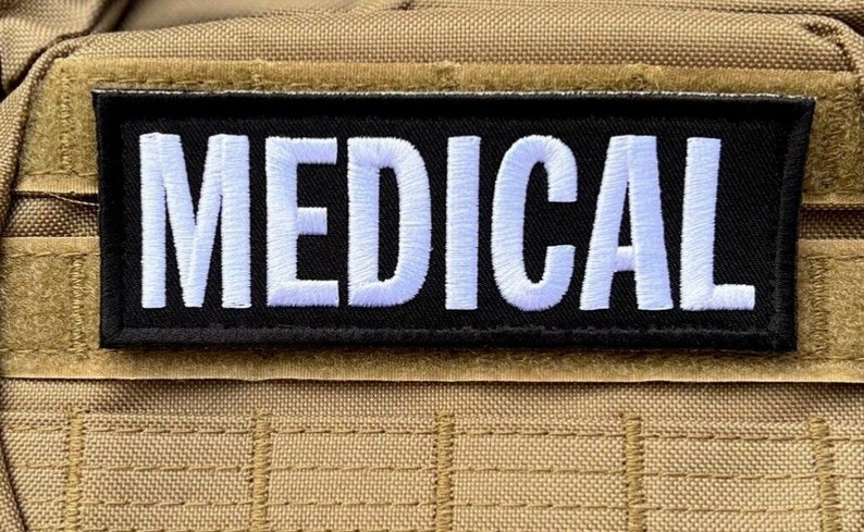 May include: A black rectangular patch with the word "MEDICAL" embroidered in white. The patch is attached to a tan fabric with a grid-like pattern, likely part of a bag or tactical gear.