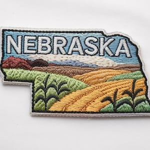May include: Embroidered patch in the shape of Nebraska, with a landscape scene. The patch shows the word "NEBRASKA" in white letters against a blue sky. The landscape includes fields of green, brown, and yellow.