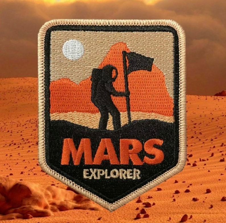 May include: A beige and black embroidered patch featuring a stylised image of an astronaut planting a flag on Mars. The patch reads "MARS EXPLORER" in orange lettering.