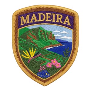 Könnte beinhalten: Ein schildförmiger gestickter Aufnäher mit dem Text MADEIRA in goldenen Buchstaben auf violettem Grund. Das Design zeigt eine grüne Berglandschaft, einen blauen Ozean, eine gelbe Paradiesvogelblume sowie kleine rosa und blaue Blüten.