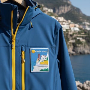 Peut inclure: Veste bleue avec fermeture &eacute;clair jaune et capuche. Elle pr&eacute;sente un &eacute;cusson avec l'inscription "AMALFI COAST" et une illustration c&ocirc;ti&egrave;re avec un phare et des b&acirc;timents. La veste est suspendue &agrave; un cintre en bois.