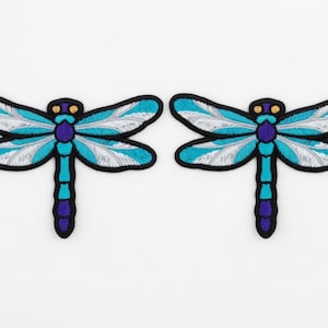 May include: Two embroidered dragonfly patches. Each patch features a black outline, teal and white wings, a purple body, and yellow accents. The patches are symmetrical and placed side by side on a white background.