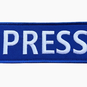 Può includere: Un cerotto rettangolare blu con testo ricamato bianco che recita "PRESS".