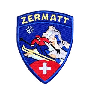 Peut inclure: Un écusson brodé bleu et jaune avec le mot "ZERMATT" en lettres jaunes. L'écusson représente un skieur en vêtements rouge et blanc, une chaîne de montagnes, un flocon de neige et un drapeau suisse.