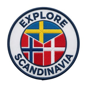 May include: A circular embroidered patch with a white background and a dark blue border. The text EXPLORE SCANDINAVIA is stitched in dark blue along the top and bottom. The centre features a triangular arrangement of the Swedish, Norwegian, and Danish flags.