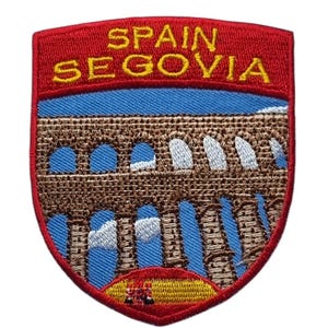 May include: Embroidered patch depicting the Segovia aqueduct in Spain. The patch has a red border with the words "SPAIN SEGOVIA" in yellow. The aqueduct is brown with blue and white details, set against a blue sky.