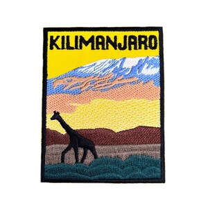 May include: Embroidered patch with a yellow background and the text "KILIMANJARO" in black. The patch depicts a black silhouette of a giraffe walking in a green field with a mountain range in the background. The sky is a gradient of pink, blue, and yellow.