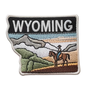 Op de afbeelding: Geborduurde patch in de vorm van Wyoming, met een landschap met bergen, een ruiter te paard en het woord "WYOMING" in witte letters op een zwarte achtergrond. De patch heeft een witte rand.