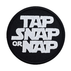 Könnte beinhalten: Ein runder, schwarzer Aufnäher mit weiß gesticktem Text "TAP SNAP OR NAP". Der Aufnäher hat einen schwarzen Rand und ist wahrscheinlich zum Aufnähen oder Aufbügeln auf Kleidung oder Accessoires gedacht.