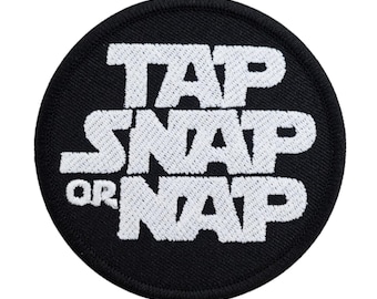May include: A round, black patch with white embroidered text that reads "TAP SNAP OR NAP". The patch has a black border and is likely designed to be sewn or ironed onto clothing or accessories.