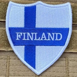 Peut inclure: Un écusson brodé blanc et bleu avec le mot "FINLAND" en lettres blanches. L'écusson a la forme d'un bouclier.