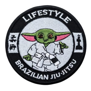 May include: A circular patch featuring a cartoon character in a white gi, holding a black belt. The patch has the words "LIFESTYLE" and "BRAZILIAN JIU-JITSU" in white text on a black background. The character has green skin and large ears.