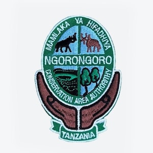 Può includere: Toppa ricamata verde e bianca con le parole "Ngorongoro Conservation Area Authority" e "Tanzania" in un cerchio. La toppa presenta un rinoceronte bianco e nero, un'antilope marrone e bianca, un albero verde e un lago blu.