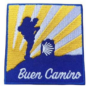 Op de afbeelding: Geborduurde patch met een blauw silhouet van een pelgrim en een sint-jakobsschelp tegen een achtergrond van gele en witte stralen. De tekst "Buen Camino" staat in witte letters op een blauwe rand.