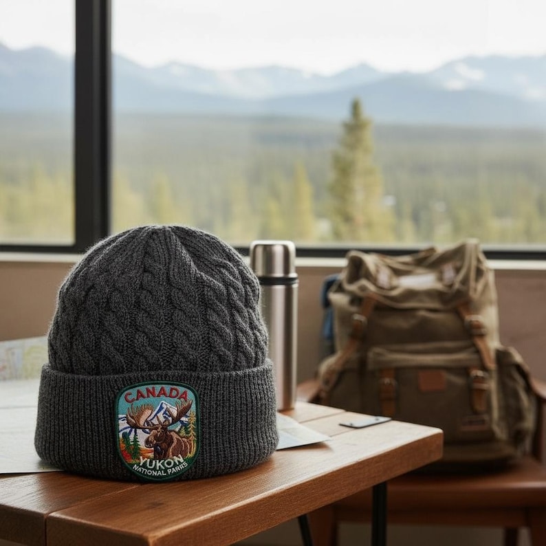 May include: A dark grey cable-knit beanie with a folded cuff. The cuff features a colourful embroidered patch depicting a moose in a mountain landscape with the text "CANADA" at the top and "YUKON NATIONAL PARKS" at the bottom of the patch.