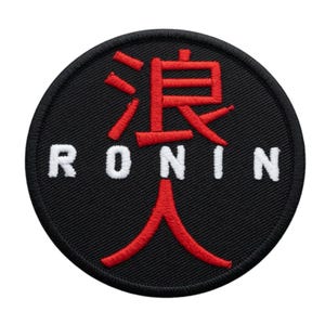 Può includere: Una toppa rotonda nera con la scritta "RONIN" in lettere bianche. Sopra la parola c'è un simbolo giapponese rosso. La toppa ha un bordo nero.