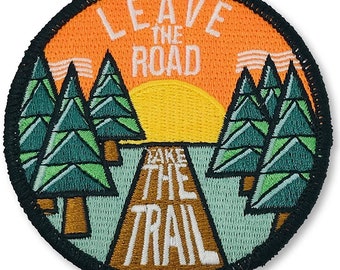 May include: A round, orange, green and brown embroidered patch with the text "Leave the Road Take the Trail" and a sunset scene with trees.