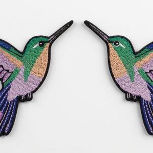 May include: Two embroidered hummingbird patches. Each patch features a hummingbird with green, blue, purple, and peach-colored details. The birds have long black beaks and are set against a white background.
