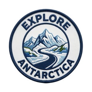 May include: A circular embroidered patch with a white background and a dark blue border. The centre features a mountain range in shades of blue and white with a winding path. The text EXPLORE ANTARCTICA is stitched in dark blue capital letters.