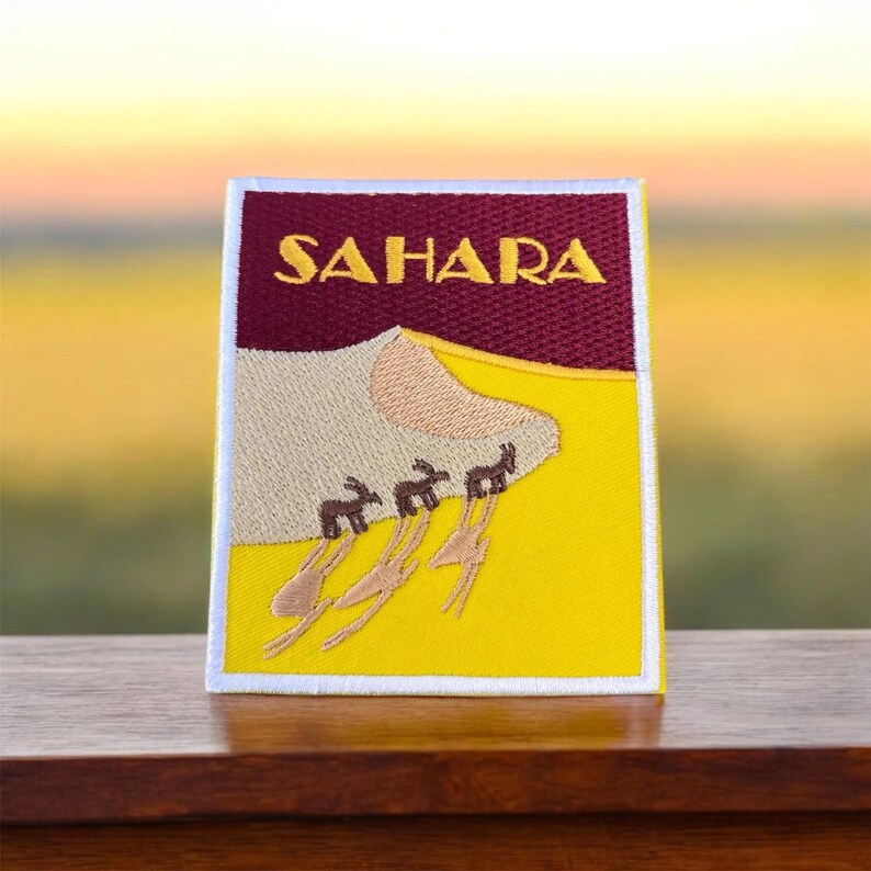 Może przedstawiać: Naszywka haftowana na ż&oacute;łto i brązowo z napisem "SAHARA" w ż&oacute;łtych literach. Naszywka przedstawia wydmę piaskową z rzędem wielbłąd&oacute;w idących po niej.
