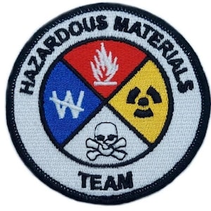 May include: A white circular patch with black text that reads "HAZARDOUS MATERIALS TEAM". The center of the patch features four colored quadrants with symbols representing different types of hazards: fire, a skull and crossbones, a radiation symbol, and a chemical symbol.